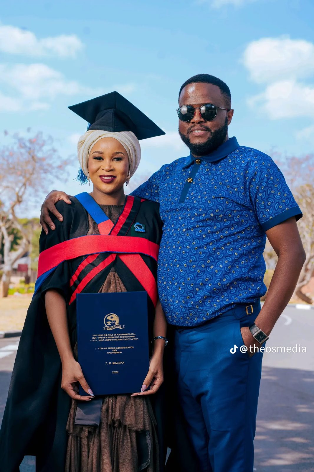 A graduate in a black gown standing with a family member outdoors