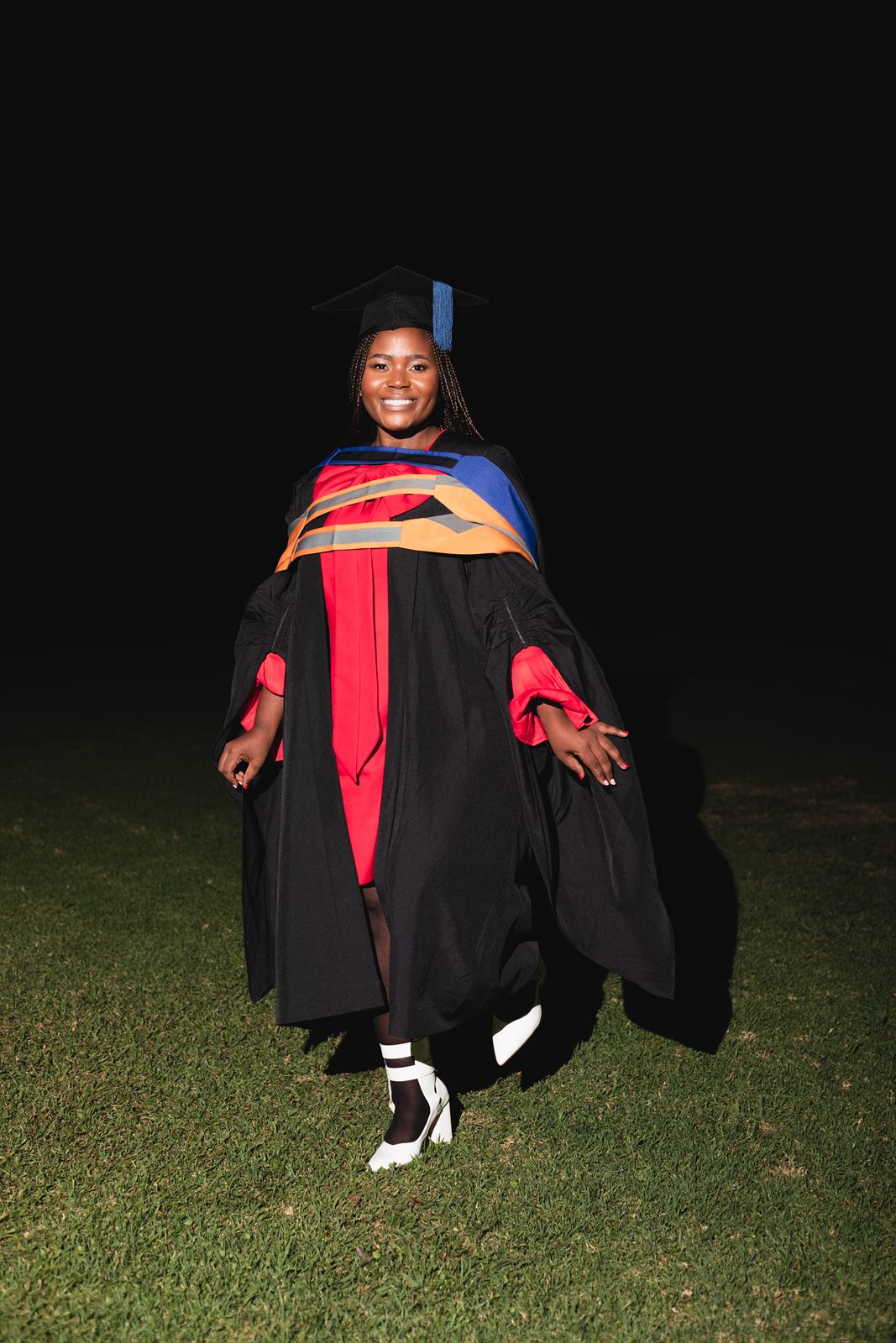 A graduate in a black gown and red dress standing on a lawn at night