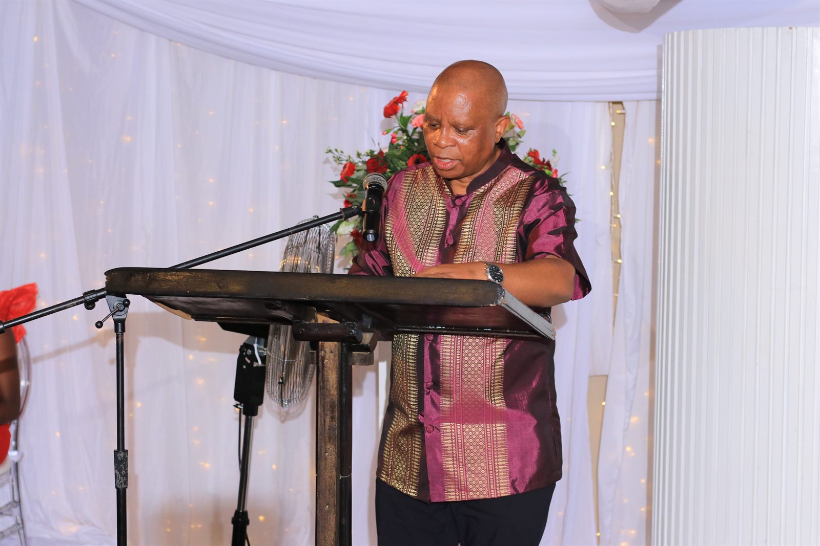 A man speaking at a podium in front of white drapes and flowers