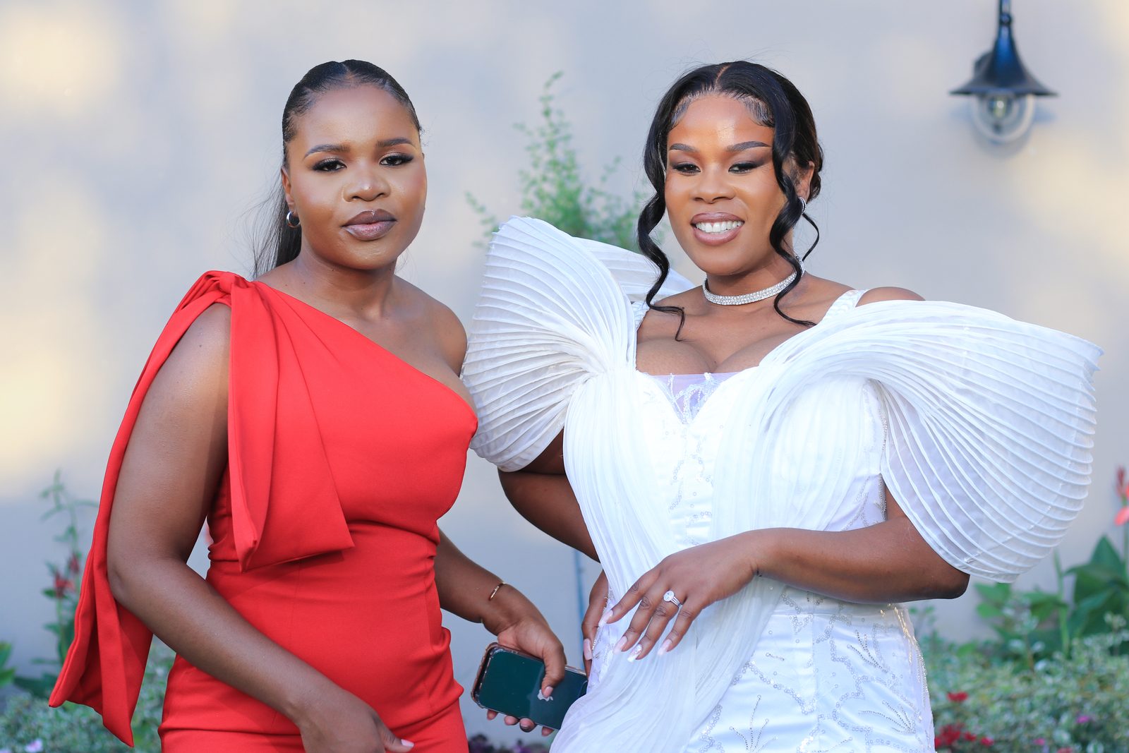 Two women posing together, one in a red dress and one in a white pleated gown