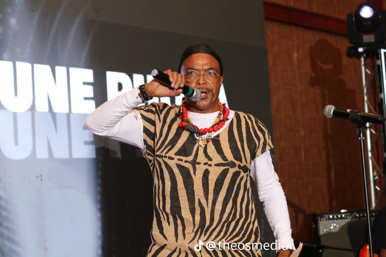A man singing into a microphone on stage in a patterned vest