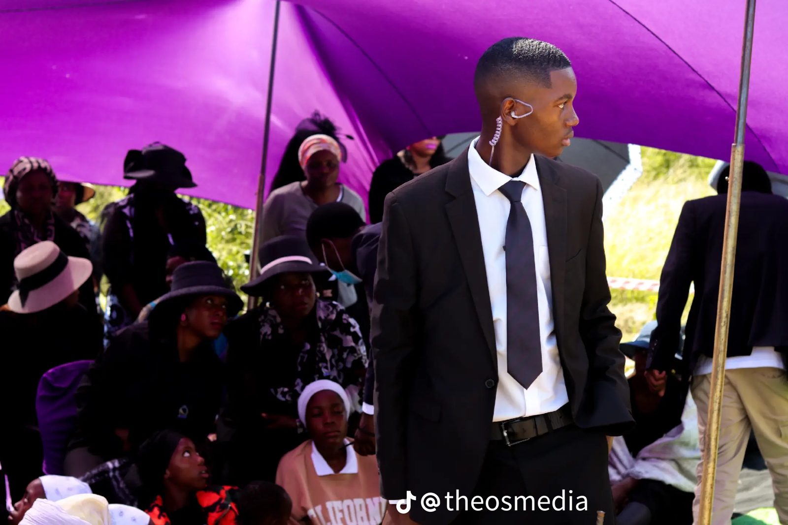 A young man in a black suit at an outdoor gathering under a purple tent