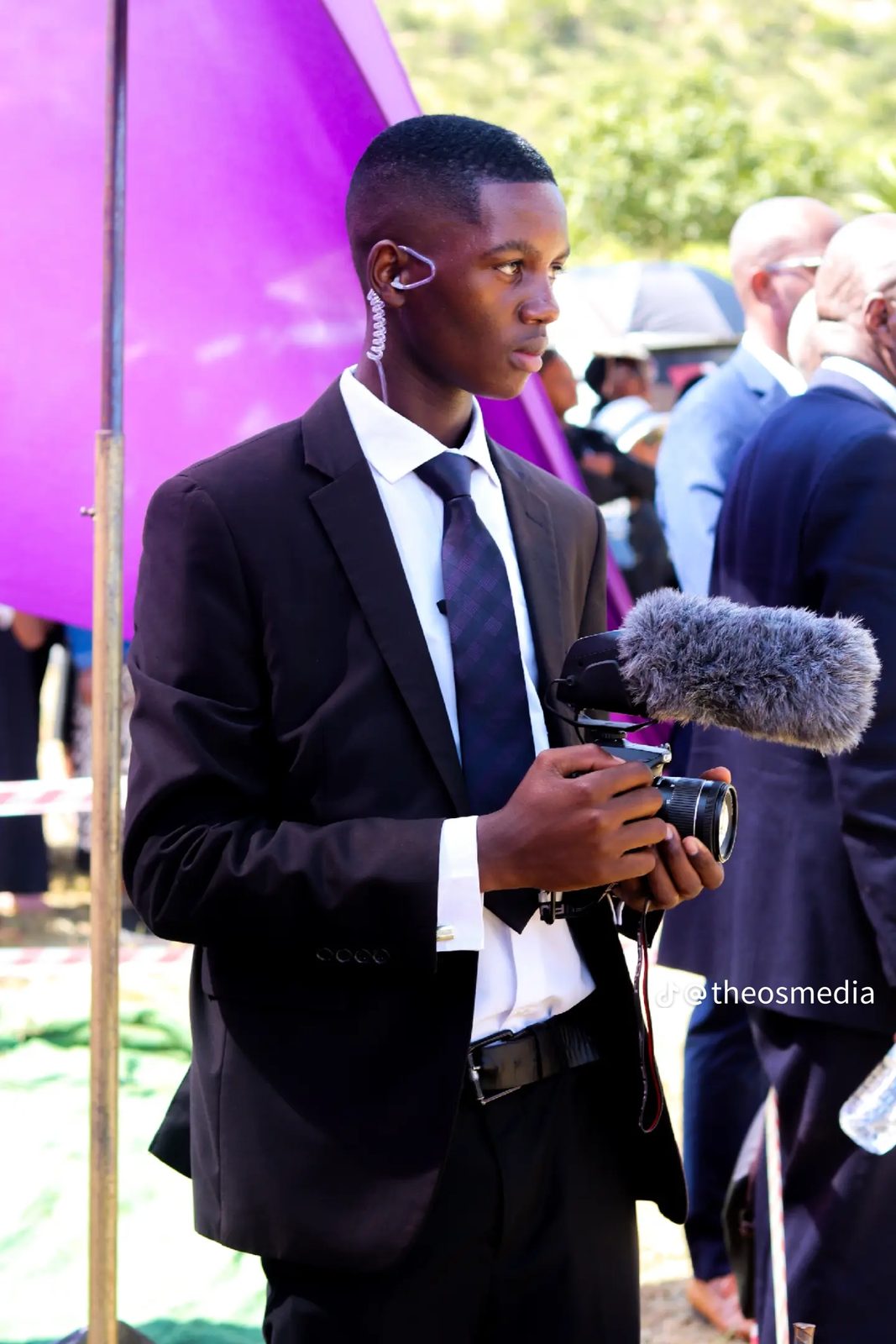 A cameraman in a black suit filming with a microphone outdoors